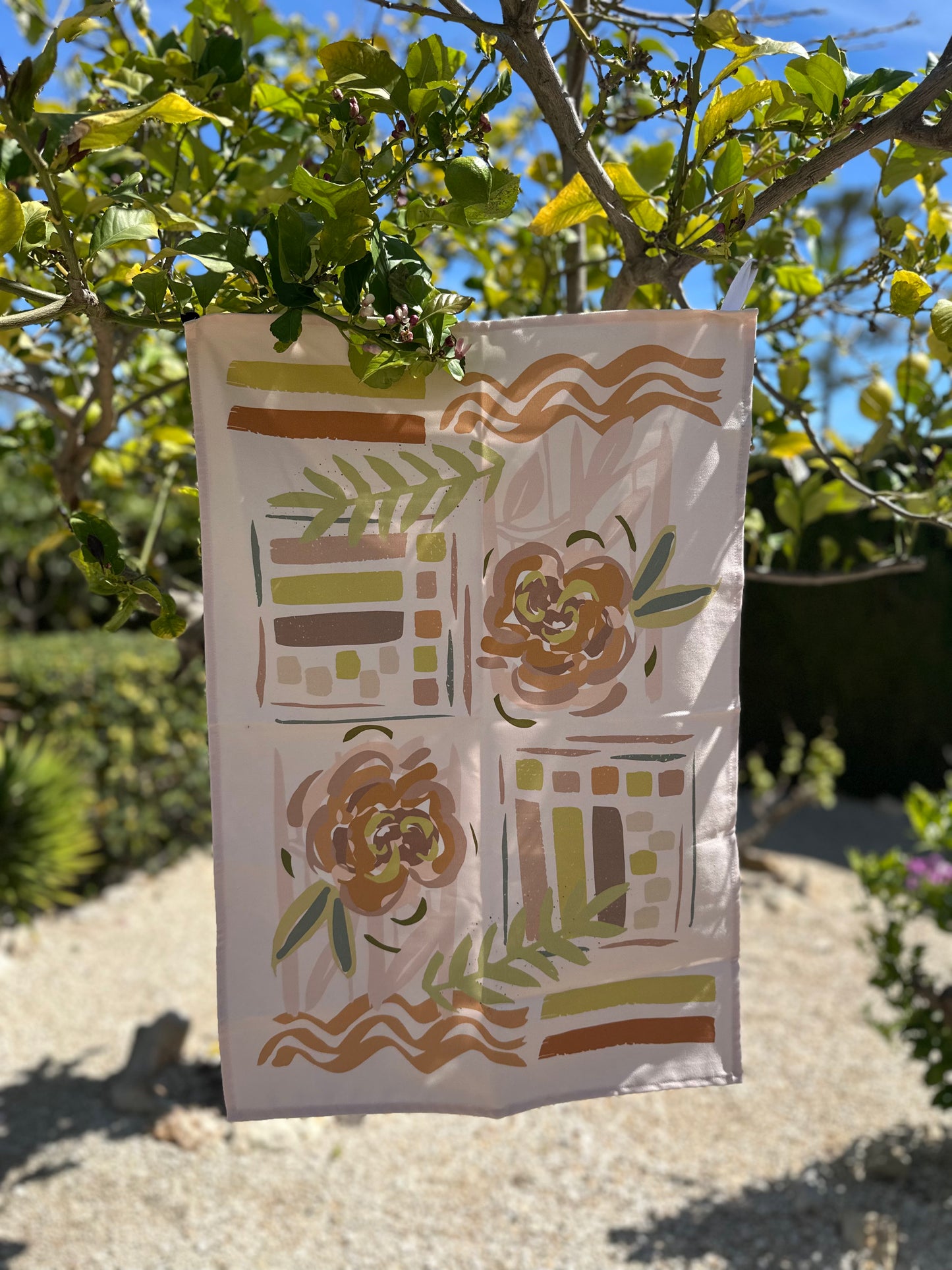 A beige kitchen towel with a large floral hand-drawn design in the center, displayed in a garden setting.