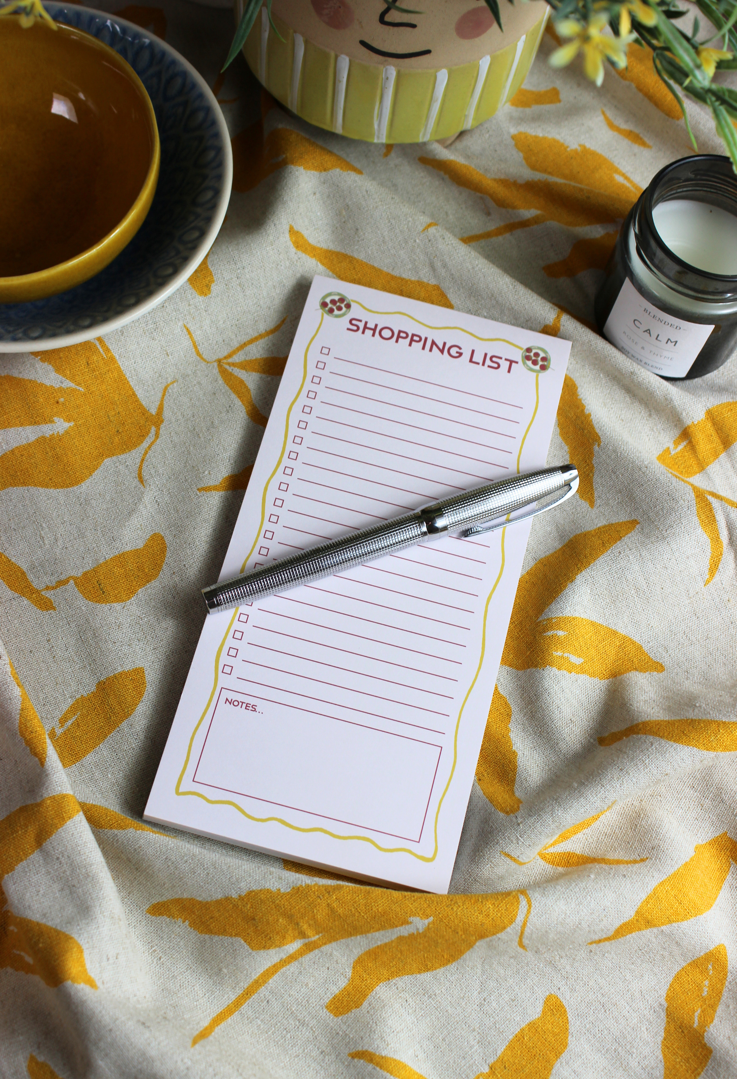 A notepad with a title 'SHOPPING LIST' on a piece of paper, placed on a table with a pen, beside a bowl and a candle, on a tablecloth with leaf patterns.