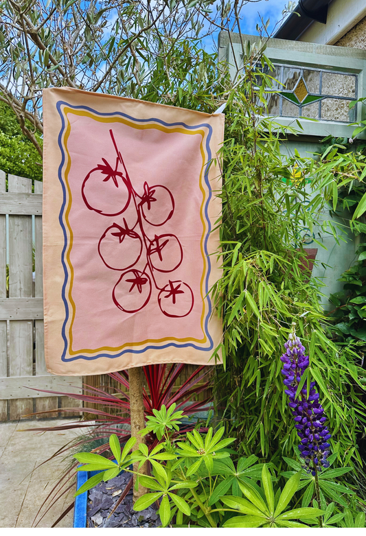 Decorative cherry tomato tea towel design on pink background hanging outdoors among plants.