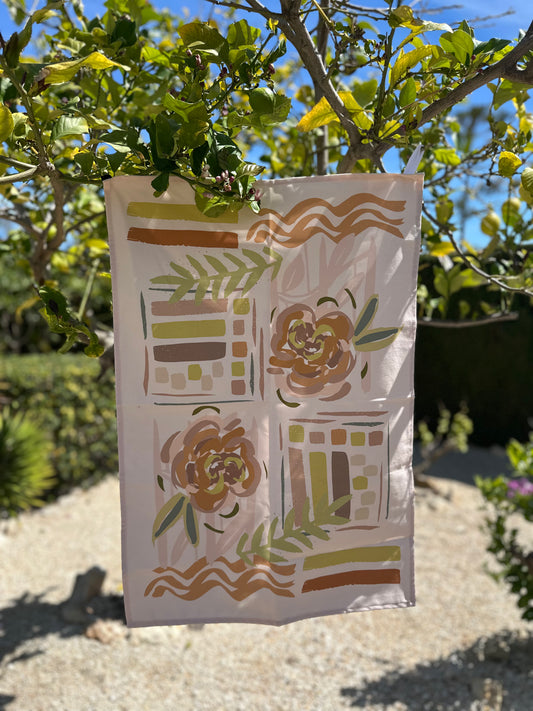 A beige kitchen towel with a large floral hand-drawn design in the center, displayed in a garden setting.
