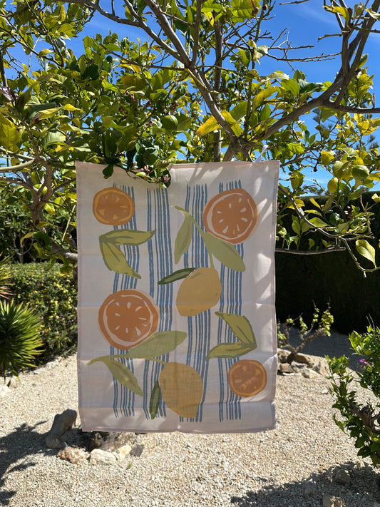 A multicolored tea towel with a design featuring oranges and lemons, displayed outdoors with foliage in the background.