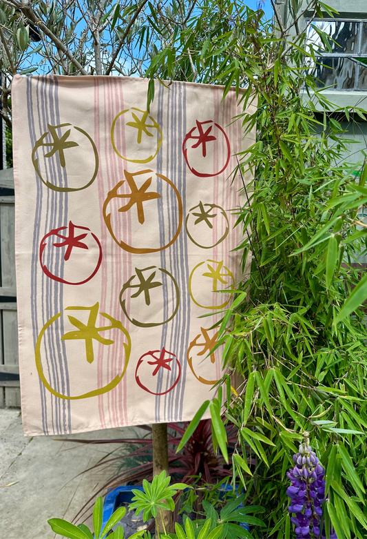 Decorative luxury tea towel with tomato hanging outdoors among plants.