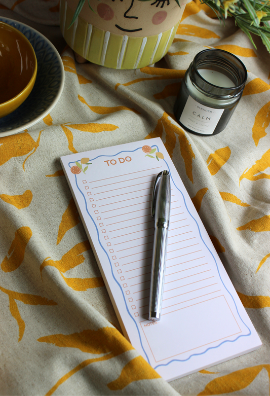 A notepad with the title 'To Do' written at the top, placed on a table with a pen on it, with a yellow and green patterned tablecloth visible.